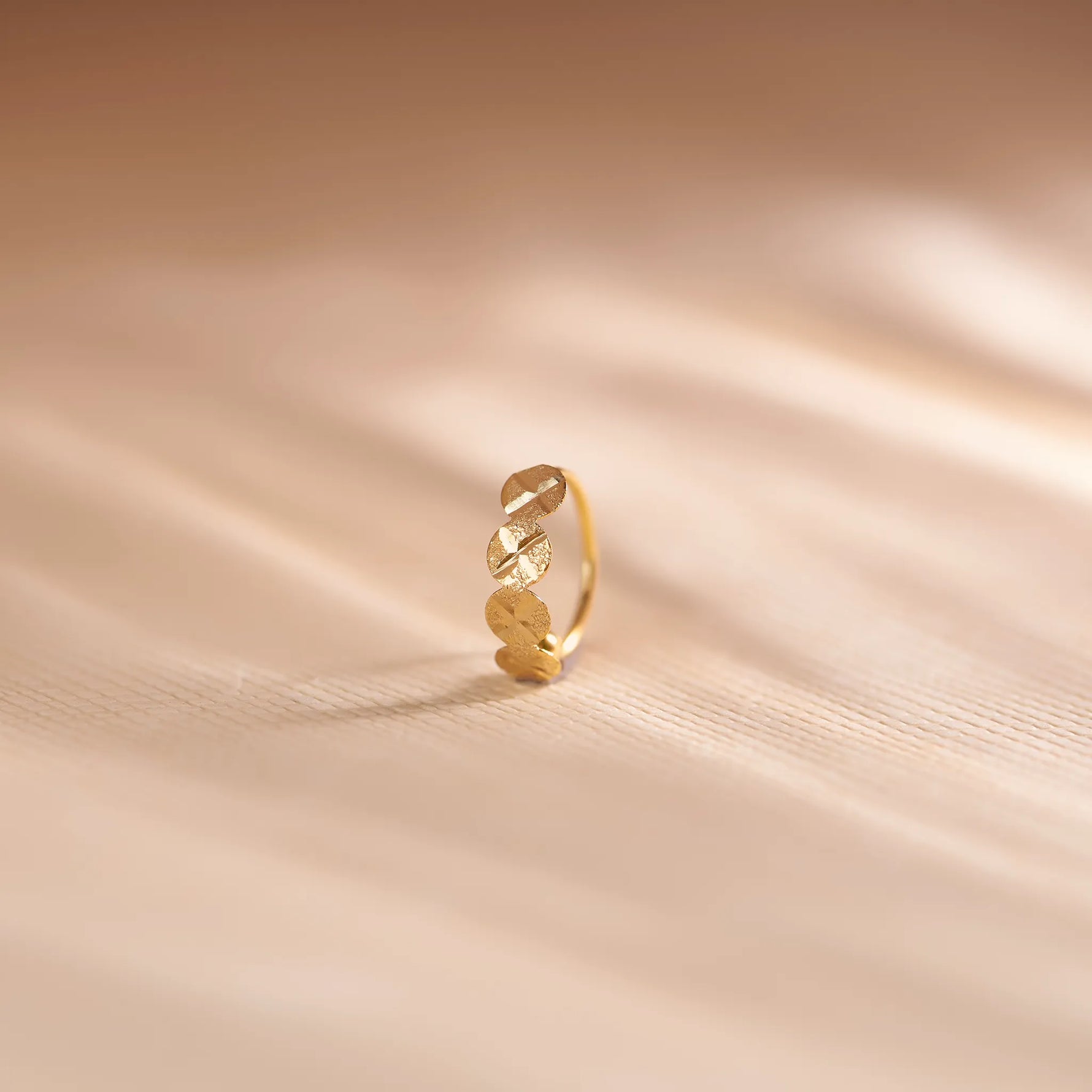 Close up of patterned 18K gold nose ring standing upright on a soft beige fabric background with gentle light waves