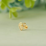 Close up of heart shaped filigree 18K gold nose pin placed on a soft pastel green surface with a blurred leafy background highlighting delicate openwork detailing