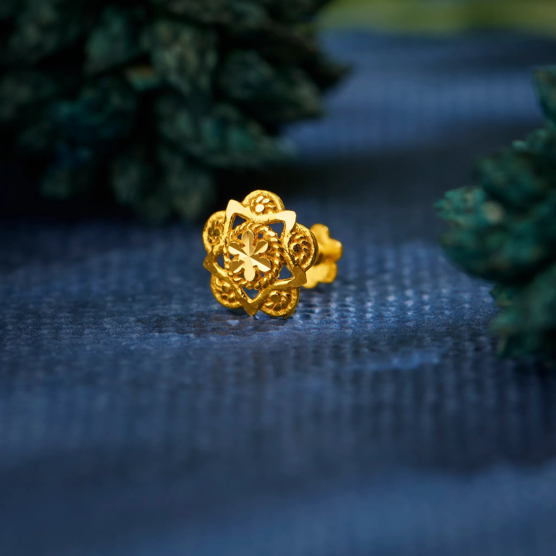 Close up view of a floral design 18K gold nose pin placed on a blue textured fabric highlighting intricate detailing