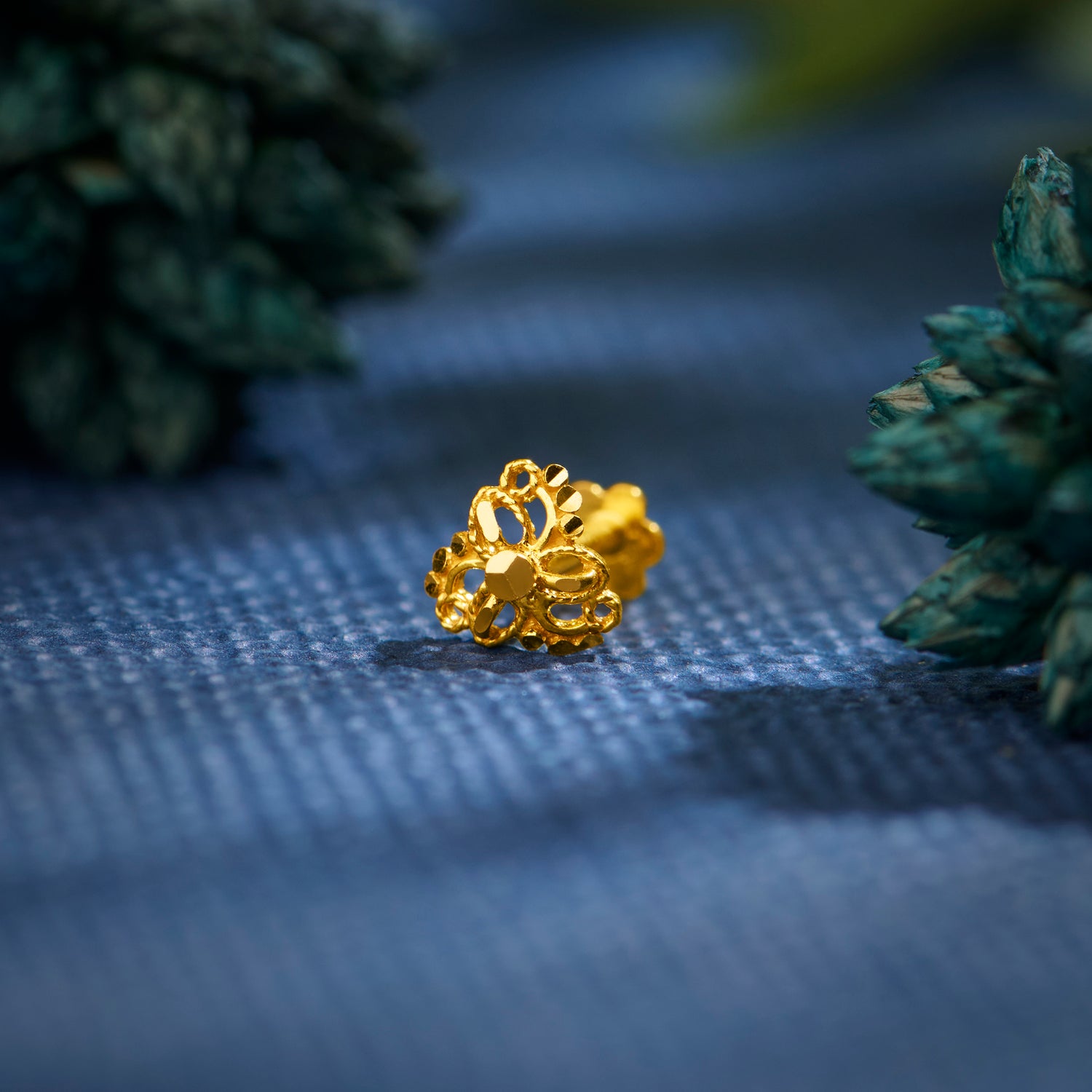 Close up of an intricate floral gold nose pin placed on a textured blue surface with greenery in the background highlighting its ornate design